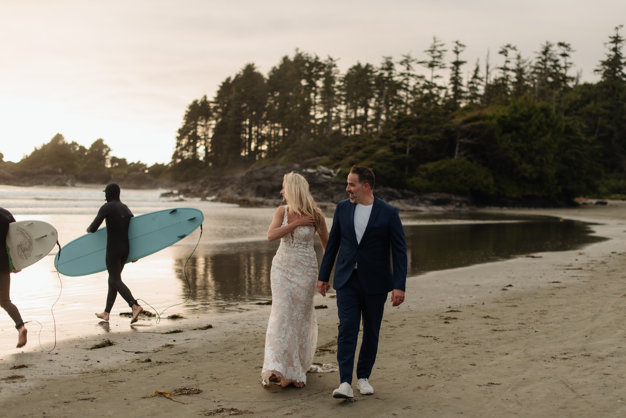 The bride and groom stroll down the beach in Tofino and encounter two surfers